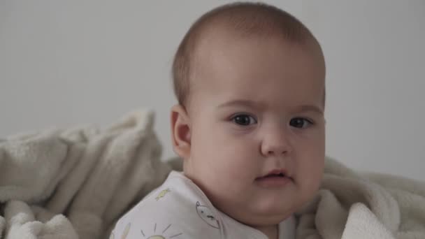 Infant, childhood concept - close-up of smiling happy funny brown-eyed chubby face of newborn kid awake. toothless 7 months baby grimaces, closes eyes sit on soft bed wrapped in warm blanket at home — Stock Video