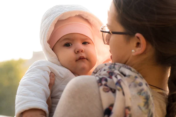 infants, childhood, motherhood, family, lifestyle concept - happy young mom in glasses hold fat chubby newborn smiling baby girl 6 months old in white jacket outdoors against backdrop of bright sun.