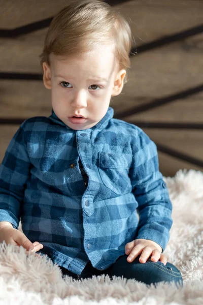 childhood, infant, minor concepts - close up one blonde black-eyed sad, offended child baby boy on soft powdery bed in sunny room. portrait embarrassed preschool Slavic kid in shirt at home indoors