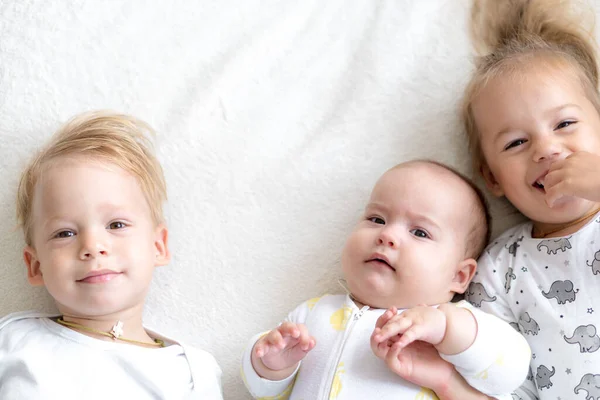 Трое маленьких улыбающихся детей играют вместе на кровати. Брат и сестра показывают новорожденному игрушку. Дети знакомятся с новорожденными братьями и сестрами. Тоддлеры смеются и сближаются. Смешные дети с небольшой разницей в возрасте. — стоковое фото