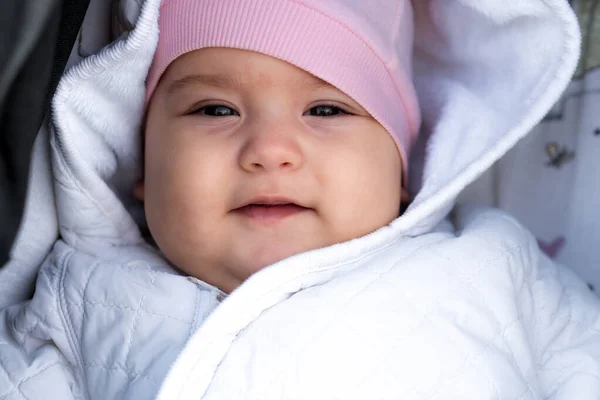 幼児,小児,感情の概念-かわいい笑顔の肖像画の顔のクローズアップ茶色目のぽっちゃり新生児目覚め歯のない赤ちゃん6-9ヶ月古いですカメラを見て横に白いジャケットピンクの帽子でベビーカー — ストック写真