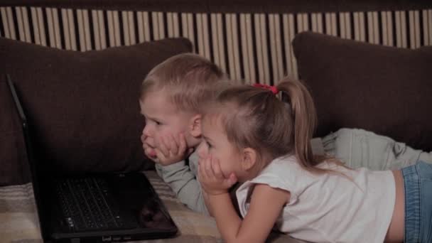 家族、ゲーム、隔離、子供時代のコンセプト-隔離中に自宅の茶色いソファの上に横たわっているラップトップで漫画を見ている2人の幸せな若い子供。兄弟兄弟姉妹は一緒に楽しみを持っている — ストック動画