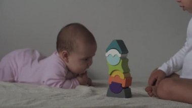 Çocukluk, aile, izolasyon, inşaat konsepti - 2 küçük mutlu çocuk yatağa oturmuş ahşap piramit inşa ediyor. 6-9 aylık yeni doğmuş kız çocuğu ve 2-3 yaşındaki oğlan evde motor becerileri için oyun oynuyorlar..