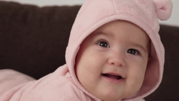 Infantil, concepto de la infancia - primer plano de sonriente feliz regordete divertido retrato de ojos marrones gordita cara de niño recién nacido despierto. muecas bebé 7 meses sin dientes, cierra los ojos se encuentran en la cama suave en rosa en la barriga. — Vídeo de stock