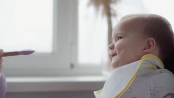 Nahrung, Ernährung, Kindheitskonzepte - Mutter füttert Säugling mit Gummilöffel. Nettes, nicht hungriges Kind in Latz gekleidet sitzt im Kinderstuhl und will in der Küche drinnen nichts essen. Abneigung gegen erste Fütterung — Stockvideo