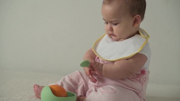 Bebeluș, copilărie, mâncare, prima hrănire, conceptul de vitamine - bucurați-vă de copil amuzant jucăuș de 8 luni fetiță îmbrăcată în îmbrăcăminte Bib mănâncă portocaliu din farfurie. dolofan copil cu lingură în interior pe fundal alb — Videoclip de stoc