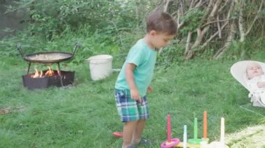 Bebeklik, çocukluk, piknik, eğitim oyunları, gelişme, yaz tatili konsepti - küçük çocuk bahçede plastik yüzük fırlatma oyunu oynuyor. Yeni doğmuş bebek sallanan sandalyede yeşil çimlerin arkasında yatıyor.