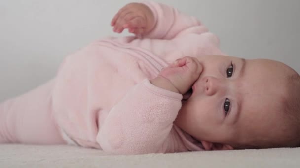 Bebé, concepto de la infancia - primer plano de sonriente feliz regordeta divertida cara gordito de ojos marrones del niño recién nacido despierto. muecas bebé 7 meses sin dientes, cierra los ojos se encuentran en la cama suave en rosa roe los dedos en la cama — Vídeo de stock