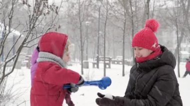 Kış, tatil, oyunlar, aile kavramları - iki mutlu anaokulu çocuğu, anneleriyle birlikte şapka ve eldiven takmış çocuklar, parkta soğuk mevsimde kartopu oynuyorlar.