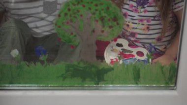 Çocukluk, yaratıcılık, doğa, ilkbahar, yaz konsepti - pencere camına akrilik renk çizen iki küçük anaokulu çocuğunun yakın çekimi. Kardeşlerin çocukları ve kız kardeşlerin manzara resimleri: ağaç, çimen