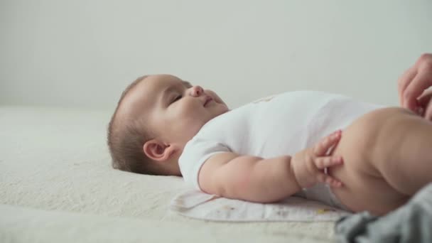 Dzieciństwo, radość, koncepcje szczęścia - słodkie maluchy aktywne dziecko grać grymaki leżące na plecach machając klaszczące ręce na białym łóżku. Mama zmienia pieluchy opatrunek pocałunek drobne dziecko w domu. — Wideo stockowe
