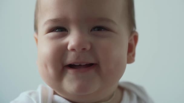 Bebé, concepto de la infancia - primer plano de sonreír feliz divertido de ojos marrones gordita cara de niño pequeño despierto. Alegría sin dientes 7 meses bebé muecas, cierra los ojos sentarse en la cama suave sobre fondo blanco en casa — Vídeo de stock
