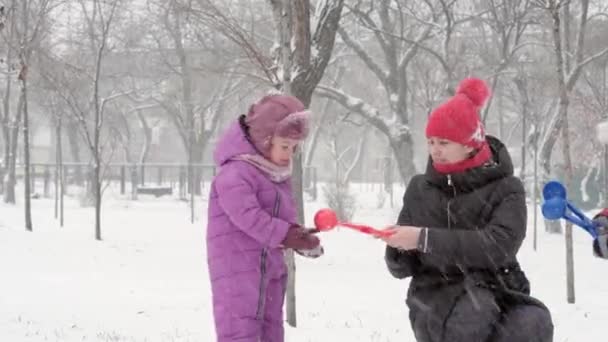Inverno, vacanza, giochi, concetti di famiglia - Due felici fratelli bambini in età prescolare vestiti con cappelli e guanti con la mamma a giocare si divertono a fare la palla di neve in nevicata nella stagione fredda nel parco — Video Stock