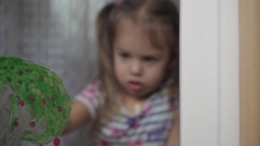 Çocukluk, yaratıcılık, doğa, ilkbahar, yaz konsepti - 3-4 yaşındaki sevimli küçük kızın yakın çekimi pencere camında akrilik renkler çiziyor. Anaokulu resim manzarası: güneş, ağaç, bulut, yağmur, çiçek