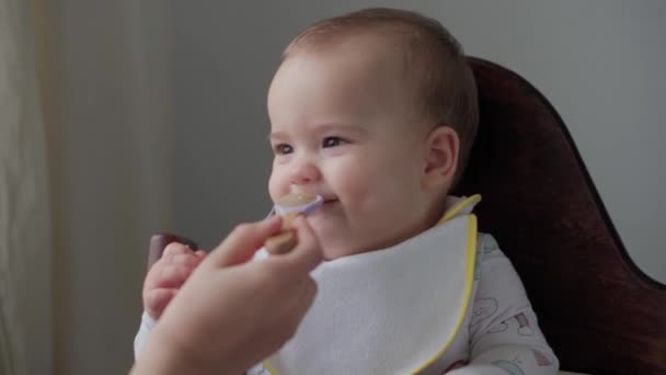 Prima alimentazione, cibo, nutrizione, concetti di infanzia - La mamma nutre il bambino con un cucchiaio di gomma. Carino bambino affamato vestito in bavaglino si siede in sedia per bambini e mangia la colazione dal piatto in cucina al chiuso — Video Stock