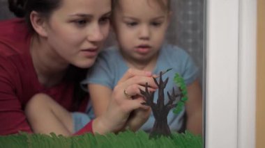 Çocukluk, annelik, yaratıcılık, doğa, sanat, bahar, yaz konsepti - 3-4 yaşlarında küçük kızı olan genç bir annenin pencere camına çizimi. kadın ve anaokulu kızı ağaç boyuyorlar..
