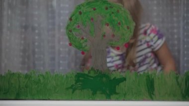 Çocukluk, yaratıcılık, doğa, ilkbahar, yaz konsepti - 3-4 yaşındaki sevimli küçük kızın yakın çekimi pencere camında akrilik renkler çiziyor. Anaokulu resim manzarası: güneş, ağaç, bulut, yağmur, çiçek