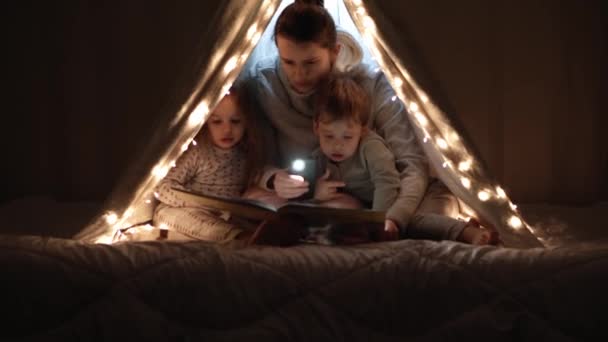 Autentikus aranyos anyuka és óvodás gyerekek fiú és lány a sátorban otthon. fiatal nő olvasni könyvet gyerekeknek 2-4 éves wigwam éjszaka. Család, gyermekkor, anyaság, kényelem és biztonság — Stock videók