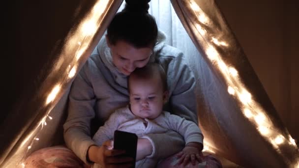 Autentikus aranyos anya pufók csecsemő lány sátorban otthon. fiatal nő használja okostelefon gyerek 6-12 hónapos ülni wigwam éjszaka. Család, gyermekkor, anyaság, kényelem és biztonság. — Stock videók