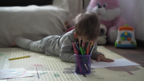 Barndom, Konst, utbildning, Kreativitet koncept - äkta förskola smart kreativ begåvad pojke 2-4 år ritar med färgade pennor bild av familjen våren sommaren, regnbåge på mattan golv hemma — Stockvideo