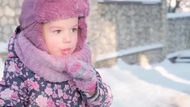Zima, wakacje, gry, koncepcje rodzinne - slo-mo zbliżenie autentyczne małe przedszkole moll 3-4 lat dziewczyna w fioletowym patrząc na aparat liże sopl z języka na śniegu. dziecko biegnie po śnieżnej okolicy — Wideo stockowe