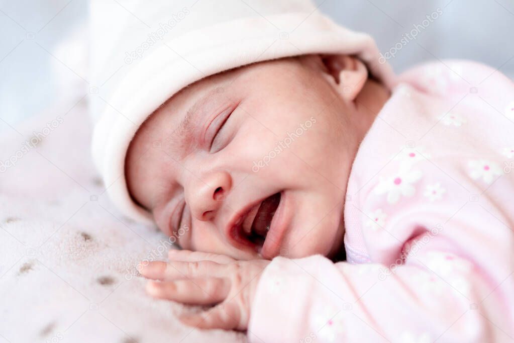 Childhood, illness, colic, bloating, motherhood, health concepts - Close up restless, worried sad newborn baby girl in hat sleeps crying scream have stomachache laying on tummy on bed with open mouth