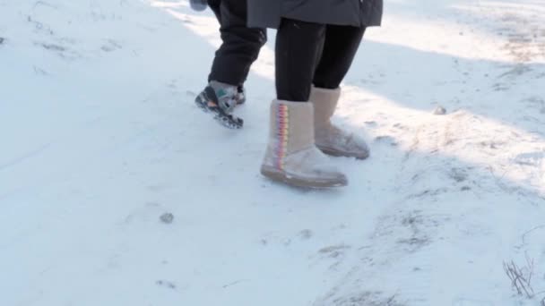 Winter, vacation, games, family concept - slo-mo close up child with mom walks through snow. feets tread on snowy path. preschool toddler kid dressed in black jumpsuit in snowfall in sunny cold season — Stock Video