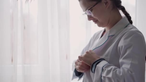 Medicina e saúde, pediatria, conceito covid-19 - close-up jovem mulher colocar o botão vestido médico branco para cima e estetoscópio contra o fundo da janela. Enfermeira ou fato médico. preparação para a cirurgia — Vídeo de Stock