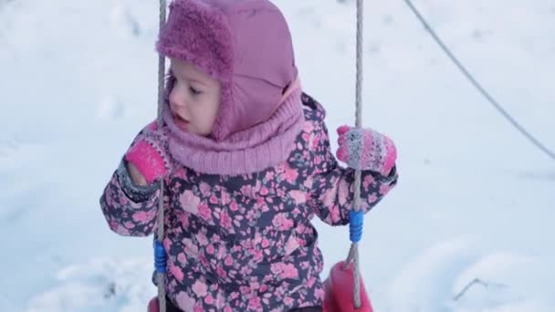 Winter, vacation, games, family concepts - middle plan of authentic little preschool minor 3-4 years old girl in purple on snow-covered meadow rides on swing before sunset. child runs on snowy area — Stock Video