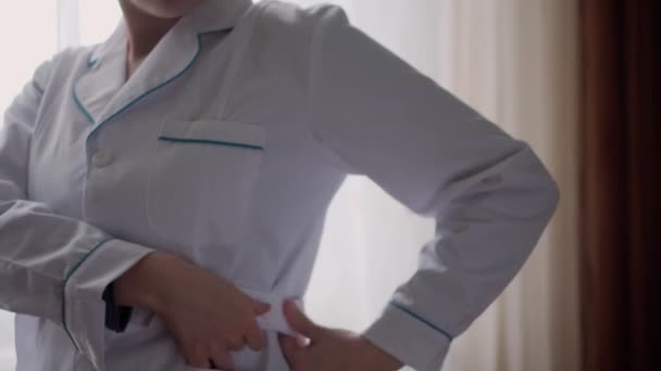 Medicina e saúde, pediatria, conceito covid-19 - close-up jovem mulher colocar o botão vestido médico branco para cima e estetoscópio contra o fundo da janela. Enfermeira ou fato médico. preparação para a cirurgia — Vídeo de Stock