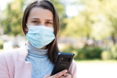 Bakım, bebekler, ilkbahar, covid-19, karantina konsepti - Genç, uzun saçlı, Avrupalı, beyaz kadın tıbbi koruyucu maskeli, gün ortasında parkın arka planında akıllı telefon kullanıyor