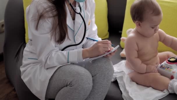 Medicina e saúde, pediatria, covid-19 conceito close-up jovem enfermeira ou médico pediatra de etnia eslava branca examina bebê bebê 8-12 meses no sofá cinza amarelo janela oposta — Vídeo de Stock