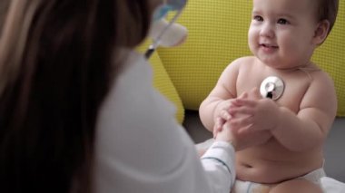 Tıp ve sağlık, pediatri, covid-19 konsepti - Kafkas Slav kökenli genç kadın hemşire ya da doktor çocuk doktorunu 8-12 aylık bebeği sarı gri kanepede muayene ediyor.