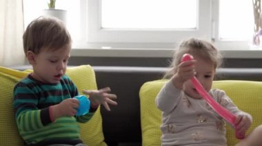 Mavi ve kırmızı sümüklü otantik bebek elleri. Kız ve oğlan anaokulunda yan yana oyun hamuru esnetiyorlar. Çocuk plastikçilik oynuyor. Modaya uygun sıvı oyuncak sarı kanepede el ve parmaklara yapışıyor.