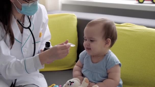 Medicina e saúde, pediatria, covid-19 conceito close-up jovem enfermeira ou médico pediatra de etnia eslava branca examina bebê bebê 8-12 meses no sofá cinza amarelo janela oposta — Vídeo de Stock