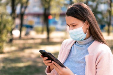 Bakım, bebekler, ilkbahar, covid-19, karantina konsepti - Genç, uzun saçlı, Avrupalı, beyaz kadın tıbbi koruyucu maskeli, gün ortasında parkın arka planında akıllı telefon kullanıyor