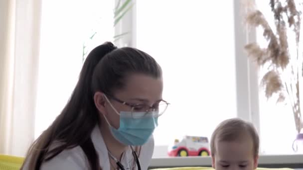 Medicina e saúde, pediatria, covid-19 conceito close-up jovem enfermeira ou médico pediatra de etnia eslava branca examina bebê bebê 8-12 meses no sofá cinza amarelo janela oposta — Vídeo de Stock