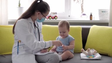 Tıp ve sağlık, pediatri, covid-19 konsepti - Kafkas Slav kökenli genç kadın hemşire ya da doktor çocuk doktorunu 8-12 aylık bebeği sarı gri kanepede muayene ediyor.