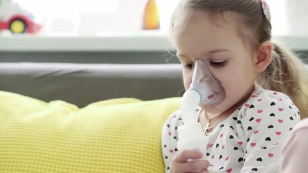 Zavřít Kavkazský 3-4 roky menší školačka dítě v masce inhalátoru na žluté šedé pohovce. Dítě užívá respirační terapii s rozprašovačem. medicína a zdraví, pediatrie, koncept covid-19 — Stock video