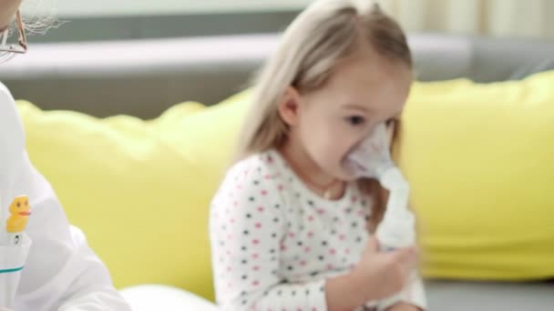 Zbliżenie młodej kobiety pielęgniarka lub lekarz pediatra sprawia, że Procedura inhalacji przez 3-4 lata dziecko przedszkolne na żółtej, szarej kanapie. Słodkie maleństwo w masce inhalatora. opiekować się chorym dzieckiem — Wideo stockowe