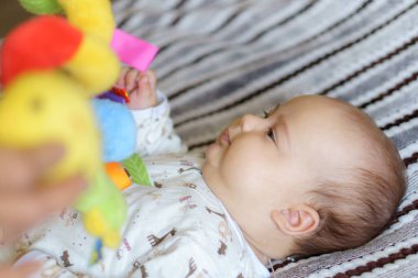 Renkli oyun minderi ve jimnastik salonunda çıngıraklı oyuncaklarla oynayan tatlı bir kız bebek. Çocuk aktivitesi ve çocuk gelişimi için oyun merkezi. Beyaz güneşli kreşte yeni doğmuş bir çocuk oyuncağı tekmeliyor ve kapıyor..