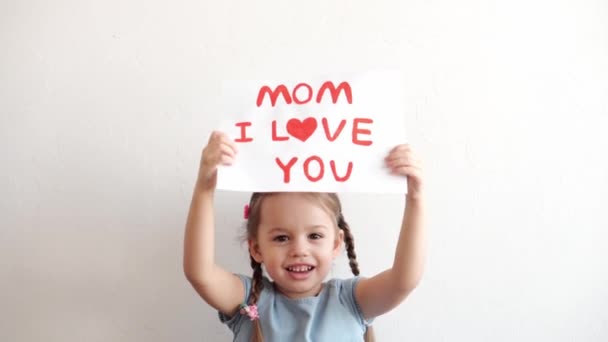 Close up cute little preschool caucasian girl 3-4 years holding white sheet with declaration of love mom. kid daughter smiling in love showing heart symbol with hands. Romantic, Mothers Day concept. — Stock Video