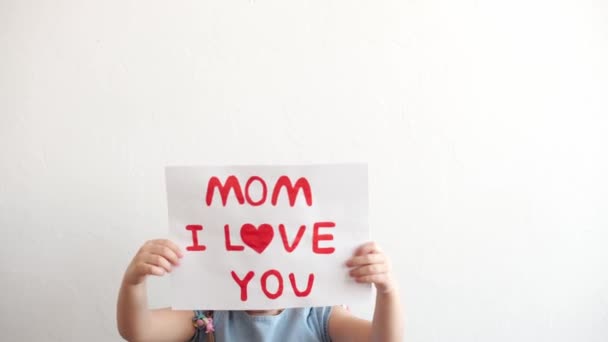 Close up cute little preschool caucasian girl 3-4 years holding white sheet with declaration of love mom. kid daughter smiling in love showing heart symbol with hands. Romantic, Mothers Day concept. — Stock Video