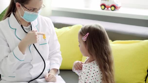Střední plán mladé bělošské slovanské ženy zdravotní sestra nebo lékař pediatr měří teplotu 3-4 roky menší školka dívka dítě na žluté šedé pohovce. medicína a zdraví, pediatrie, koncept covid-19 — Stock video