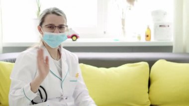 Kafkas Slav kökenli genç kadın hemşire ya da doktor çocuk doktorunun orta dereceli planı, sarı gri kanepede 2-3 yaşındaki küçük kız çocuğu muayene ediyor. ilaç ve sağlık, pediatri, covid-19 konsepti