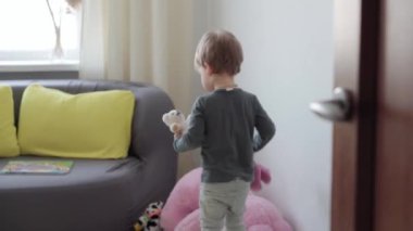 2-3 yıl süveter ve kot pantolon giyen orta sınıf otantik çocuk çocuk çocuk oyuncak ayıları çocuk odasında pencerenin ve sarı-gri kanepenin arkasına fırlatıyor. çocukluk, hayat, özgünlük kavramı