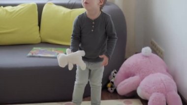 2-3 yıl süveter ve kot pantolon giyen orta sınıf otantik çocuk çocuk çocuk oyuncak ayıları çocuk odasında pencerenin ve sarı-gri kanepenin arkasına fırlatıyor. çocukluk, hayat, özgünlük kavramı