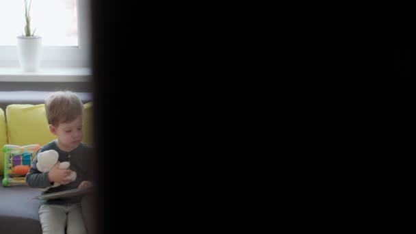 Authentische neokaukasische Brillenmutter mit 2 Kindern auf grauem, gelbem Sofa und Lesebuch. Junge Frau streichelt Säugling. Mutterschaft, Elternschaft, Kindheit, Lebenskonzept — Stockvideo