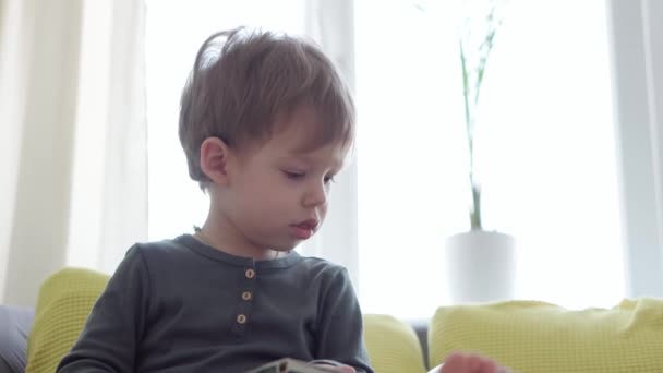 Padella verticale primo piano autentico malato bambino bambino bambino eccitato 2-3 anni esamina e legge libro con la bocca aperta finestra opposta sul divano giallo-grigio. infanzia, vita, concetto di medicina — Video Stock