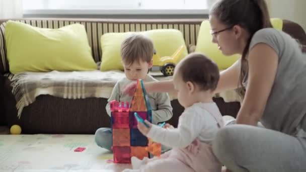 Детство, семья, материнство, материнство - молодая счастливая мать любит играть магниты блоки строит замок с малышами. Трое детей веселятся вместе в игровой комнате на карантине — стоковое видео